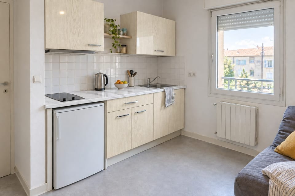 Kitchenette de studio, façades en chêne pâle, plan de travail effet marbre blanc, petit réfrigérateur sous le plan. Façades Pale Oak AG13, plan de travail Statuary White NE31. Image d'ambiance aux teintes du catalogue.