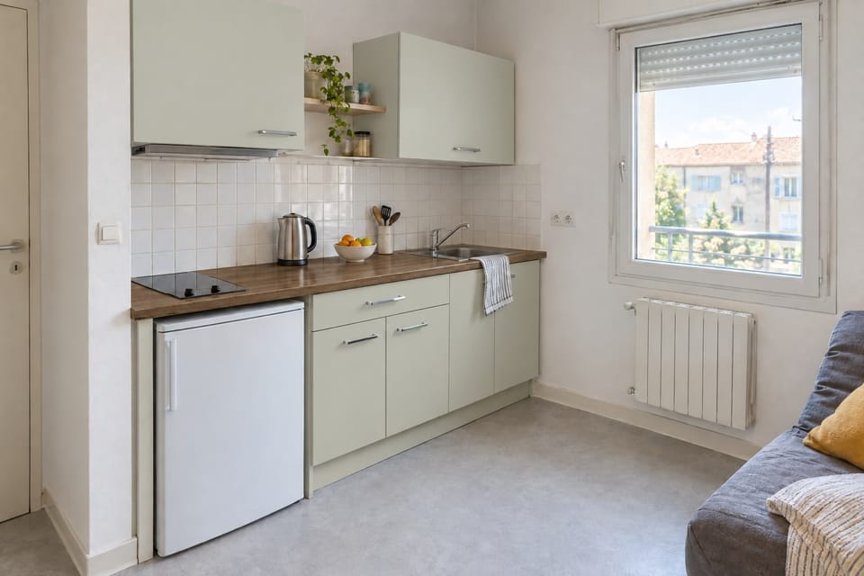 Kitchenette de studio, façades vert tendre, plan de travail en chêne, petit réfrigérateur sous le plan. Façades Soft Green NH21, plan de travail Original Oak AA14. Image d'ambiance aux teintes du catalogue.