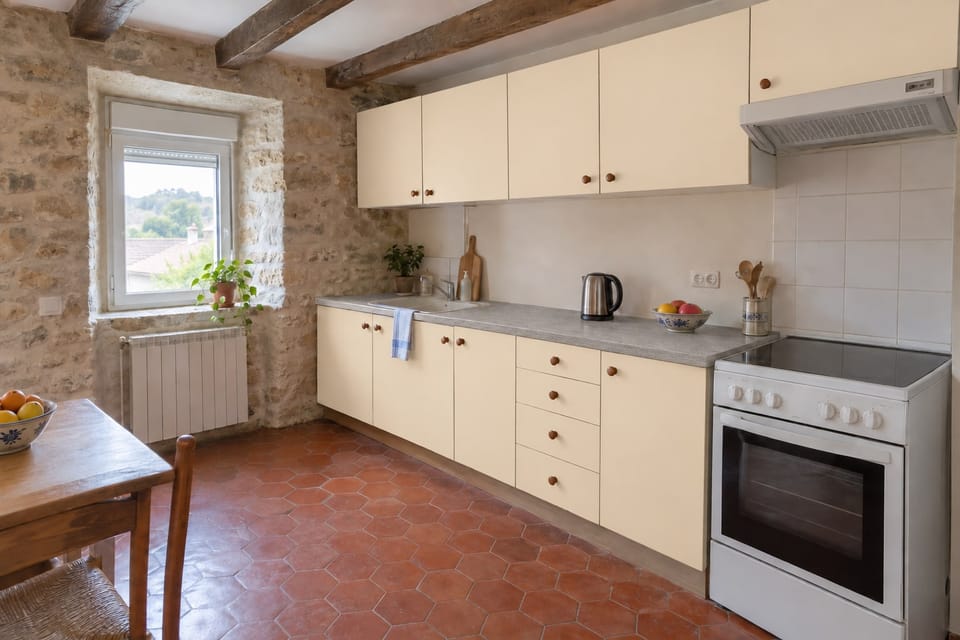 Cuisine d'une maison de village en pierre, façades crème, plan de travail effet béton gris, tomettes et poutres apparentes. Façades Oat Milk M10, plan de travail Raw Grey NE24. Image d'ambiance aux teintes du catalogue.