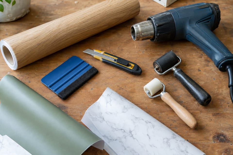 Outils de pose sur un établi : raclette feutrée, cutter, décapeur thermique, rouleaux, un rouleau de film effet chêne et des chutes vert sauge et marbre blanc. Image d'ambiance.