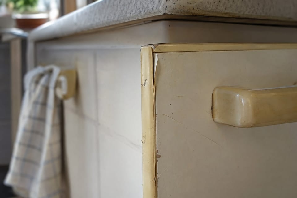 Gros plan d'une porte de cuisine blanche jaunie par les années, le chant qui se décolle, de petits éclats et une poignée en plastique. Image d'ambiance.